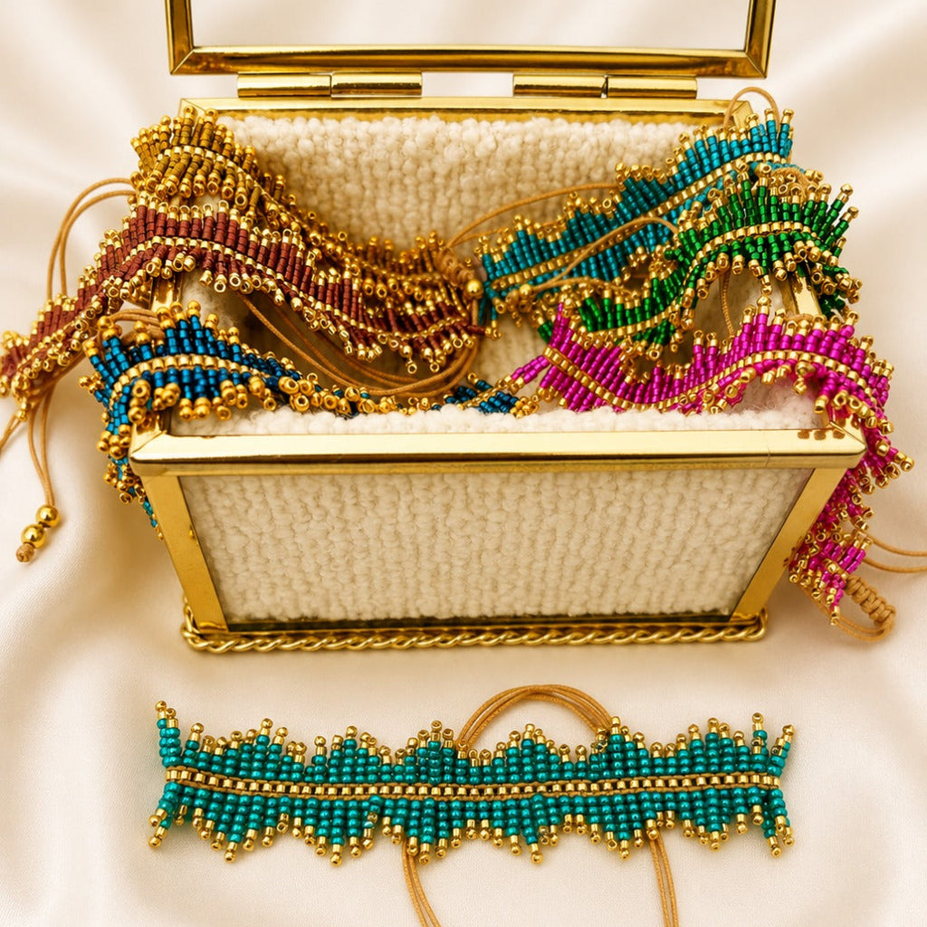 A gold-framed woven box filled with colorful beaded accessories, including turquoise, green, pink, and red woven strands with gold beads, displayed on a cream satin background. A matching turquoise and gold beaded band is laid in front of the box.