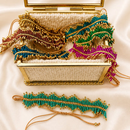 A gold-framed clutch bag with a white beaded interior is open on a cream satin background, displaying several matching beaded wristlet and anklet-style accessories in green, blue, pink, and purple with gold trim.