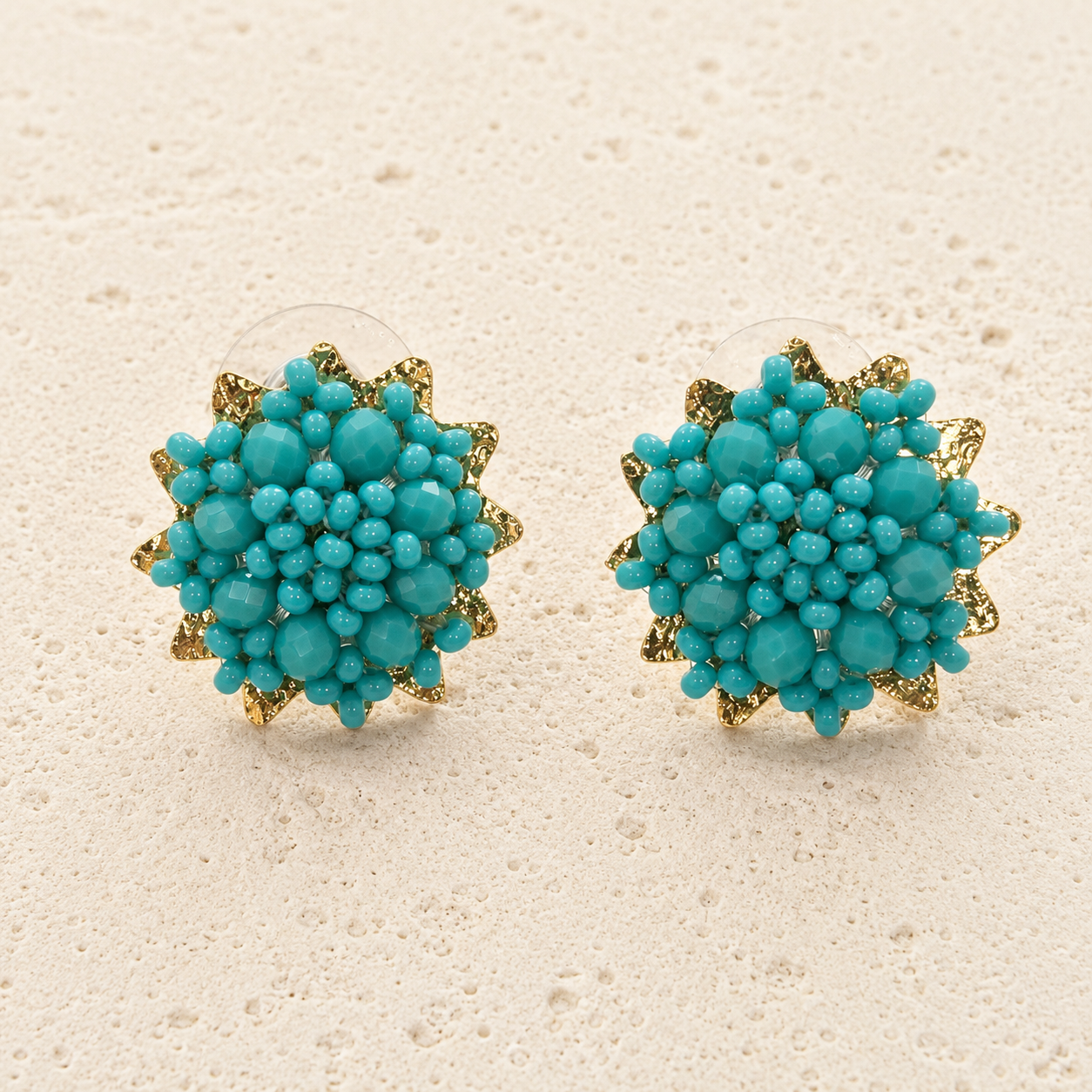 A pair of gold and turquoise floral stud earrings displayed on a textured beige surface.