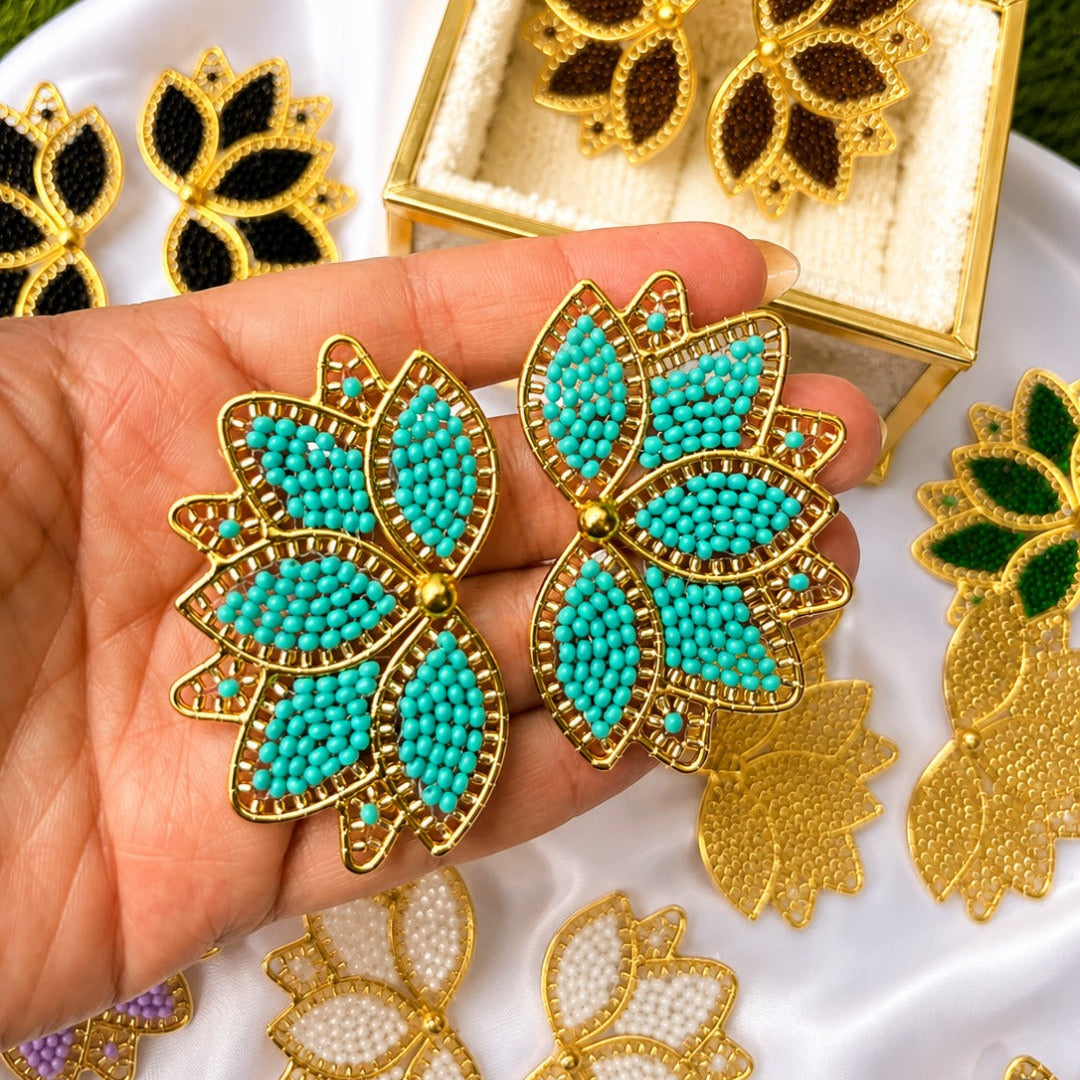 A hand holds a pair of gold-tone leaf-shaped earrings with turquoise bead centers and small red accents, with more matching earrings displayed in a gold box and around on white fabric and green grass.