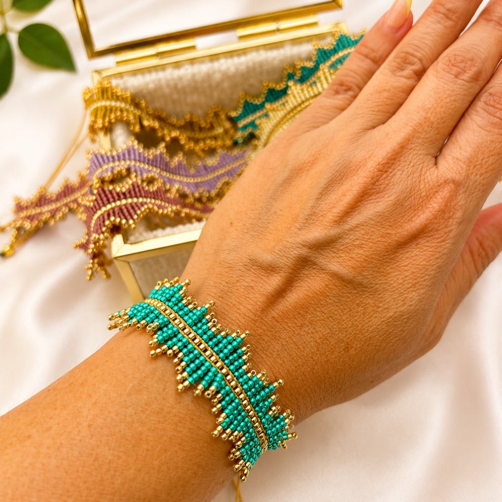 A close-up of a person's hand and wrist wearing a turquoise and gold beaded bracelet, with additional matching beaded jewelry and a gold mirror in the background on a white satin surface.
