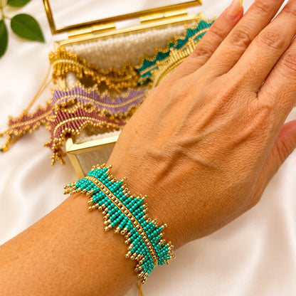 A close-up of a person's hand and wrist wearing a turquoise and gold beaded bracelet, with additional matching beaded jewelry and a gold mirror in the background on a white satin surface.