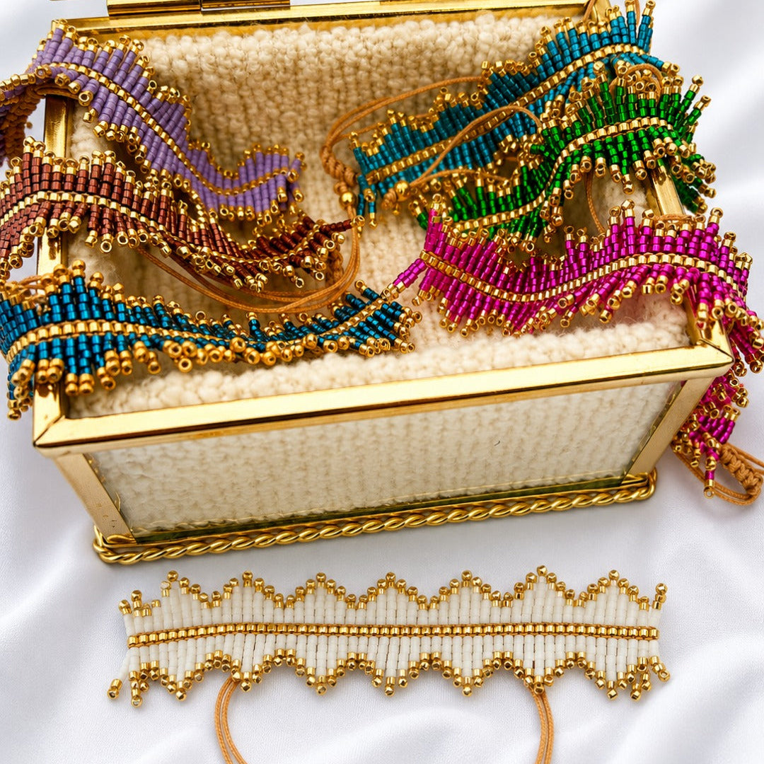 A gold-framed jewelry box filled with colorful beaded accessories, including purple, teal, green, and magenta ribbons, with a matching gold beaded choker laid out in front on a white satin background.