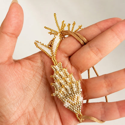 A gold and white beaded shell-shaped pendant or brooch held in a person's hand against a plain light background.