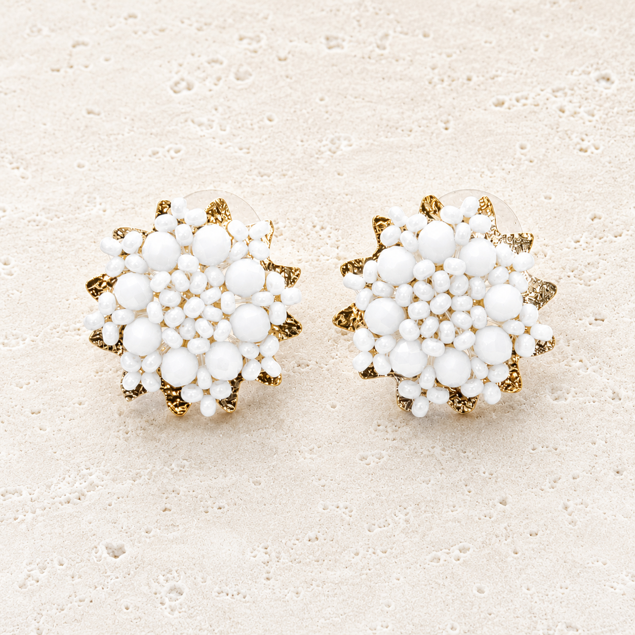 A pair of round stud earrings featuring a dense cluster of white faux pearls surrounded by textured gold-tone metal petals, displayed on a light beige textured surface.