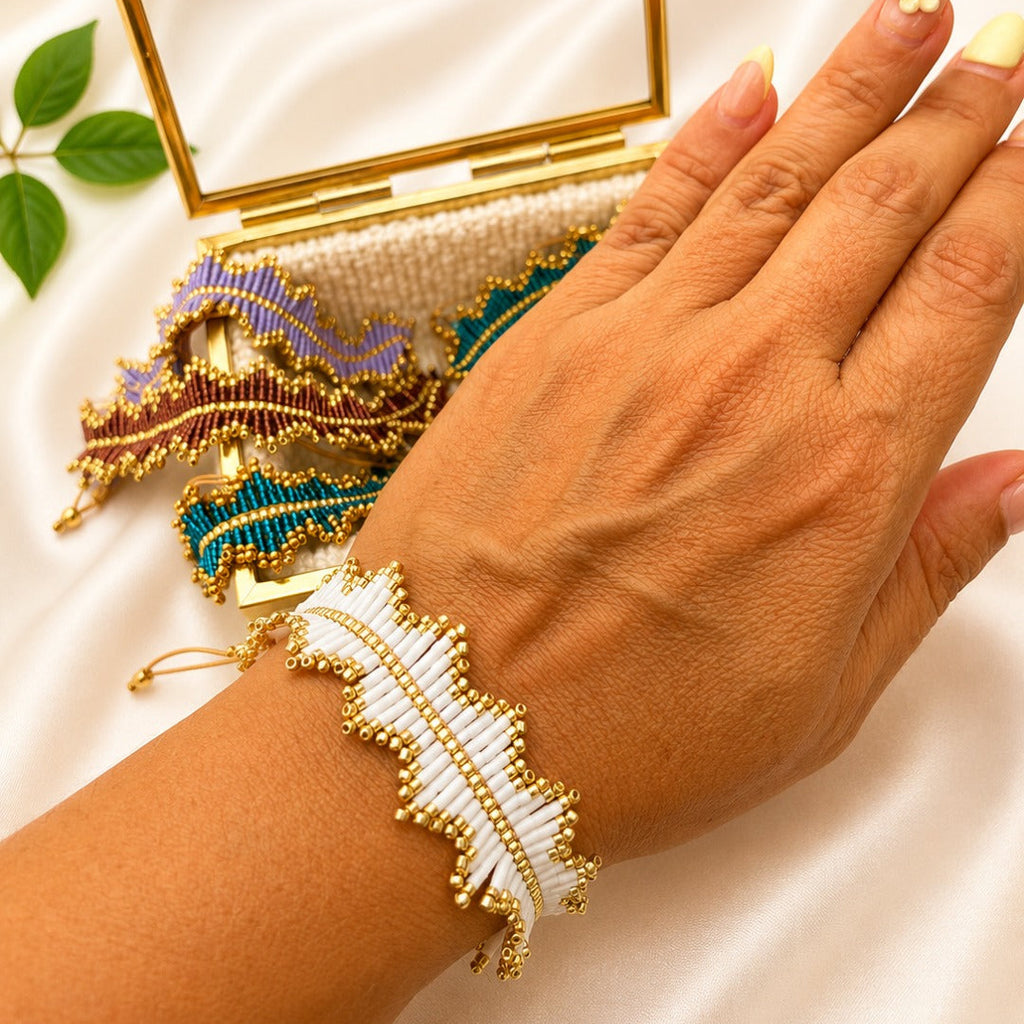 A close-up of a hand wearing a white and gold beaded feather-shaped bracelet, with a matching gold clutch case and colorful beaded accessories on a white satin background.