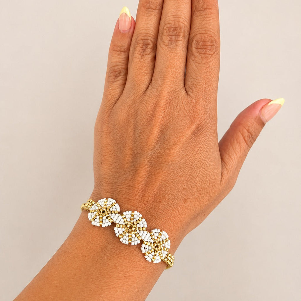 A gold-toned beaded bracelet with three flower clusters of white beads, shown worn on a person's wrist with a neutral beige background.