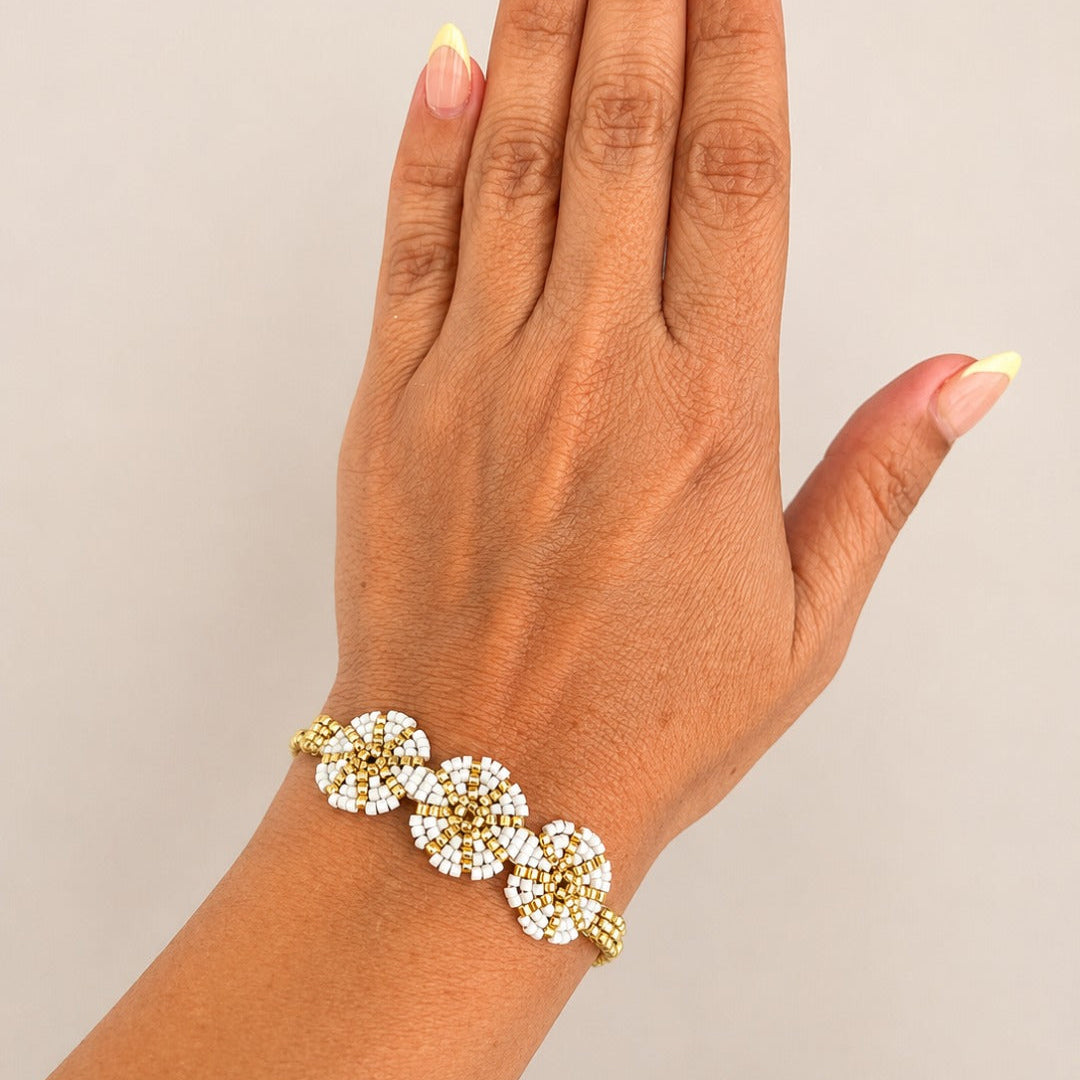 A gold-toned beaded bracelet with three flower clusters of white beads, shown worn on a person's wrist with a neutral beige background.