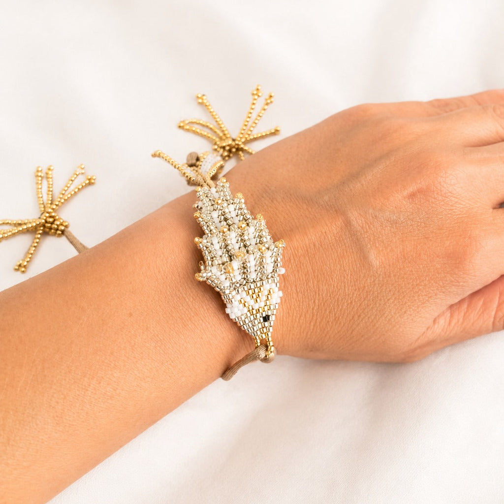 A gold-toned beaded bracelet with a large shell-shaped crystal center, shown worn on a person's wrist against a soft white fabric background.