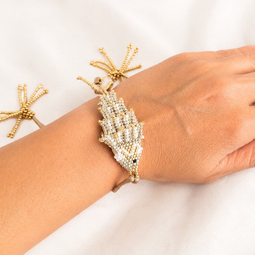 A gold-toned beaded bracelet with a large shell-shaped crystal center, shown worn on a person's wrist against a soft white fabric background.