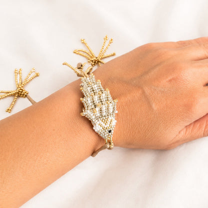 A gold-toned beaded bracelet with a large shell-shaped crystal center, shown worn on a person's wrist against a soft white fabric background.