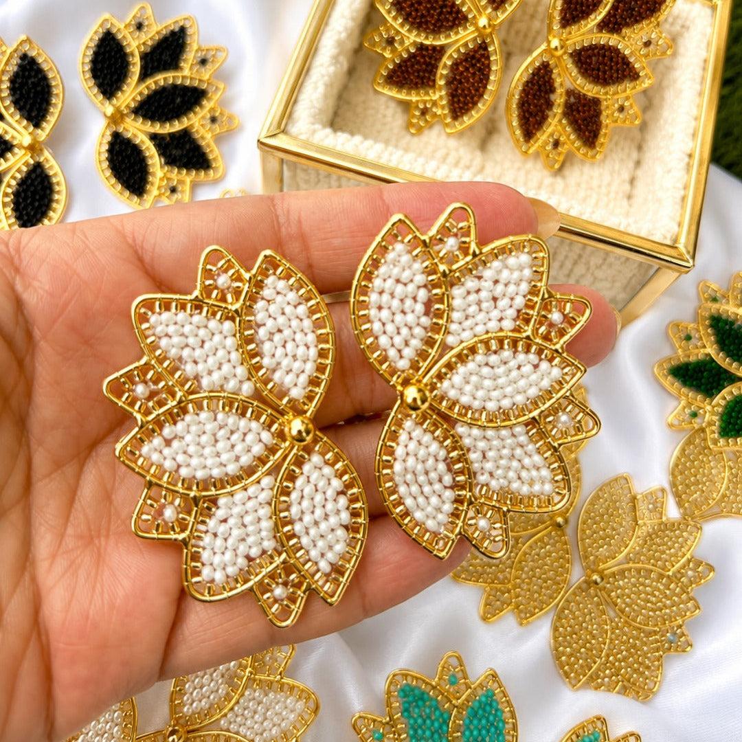 Gold-tone leaf-shaped earrings with white beadwork and small clear stones, held in a hand. A matching open jewelry box with similar leaf earrings sits behind, along with other color variants on white fabric and green grass in the background.