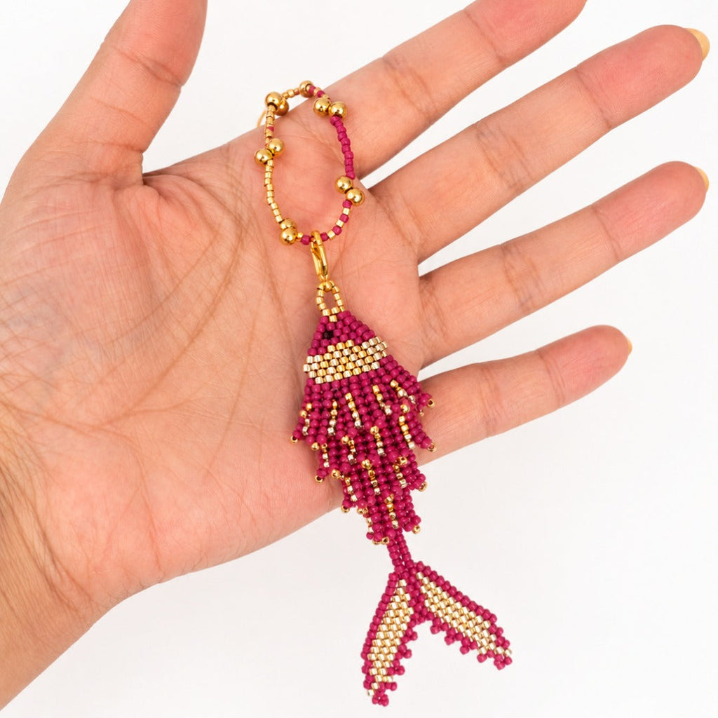 A hand holding a beaded fish-shaped keychain or bag charm in deep pink and gold seed beads against a plain white background.