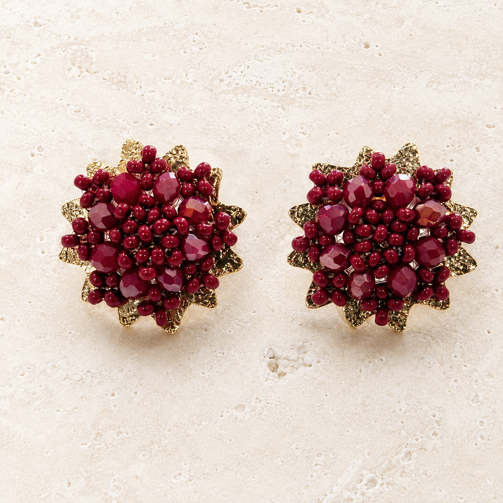 A pair of round gold-toned stud earrings decorated with clusters of faceted deep red gemstones and small red beads, displayed on a light beige textured surface.